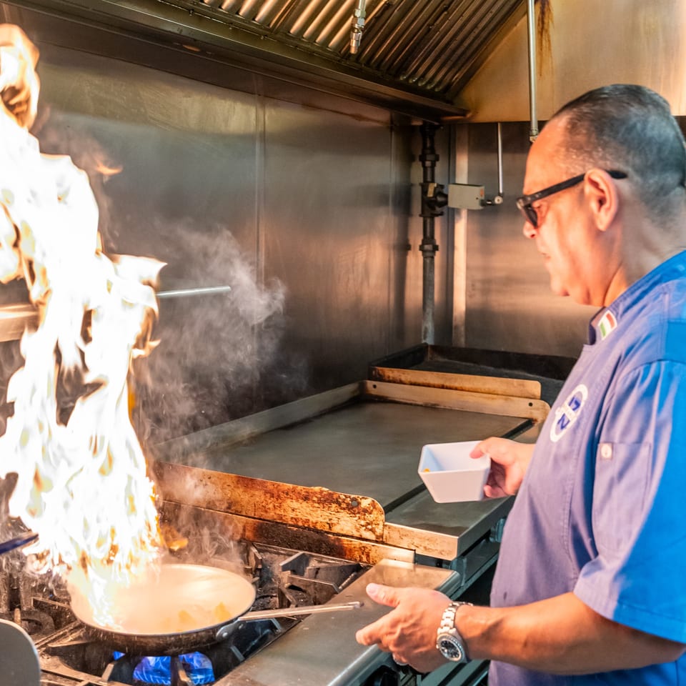 A chef in a blue shirt flambés food in a pan with large flames at Oceans 13.