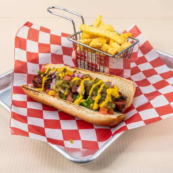 Grilled hot dog with relish, onions, tomatoes, and mustard, served with fries in a metal basket.