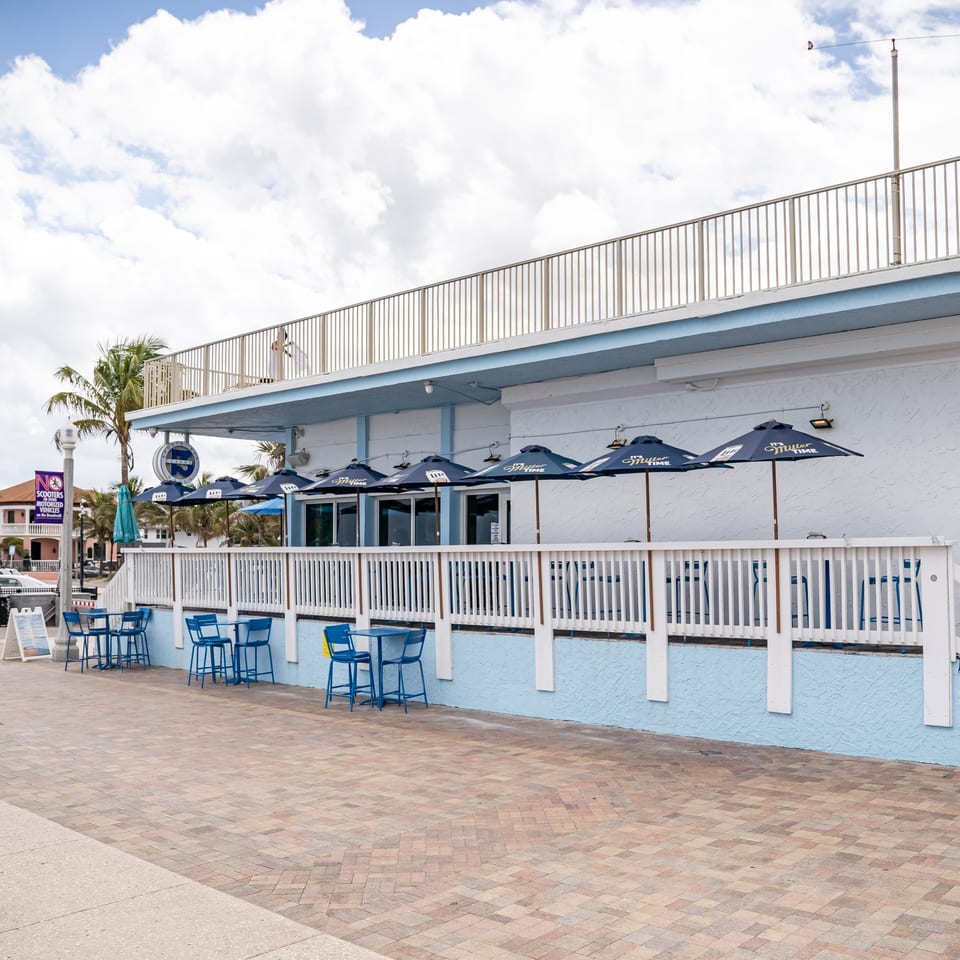 Outdoor patio of a beachside restaurant with blue tables, chairs, and umbrellas under a cloudy sky.