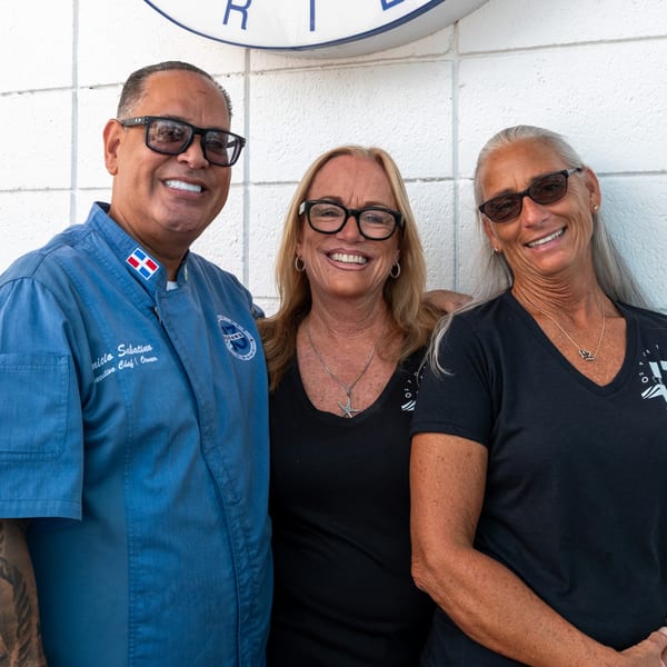 Oceans 13 chef and two women smiling, standing against a white wall with a logo.