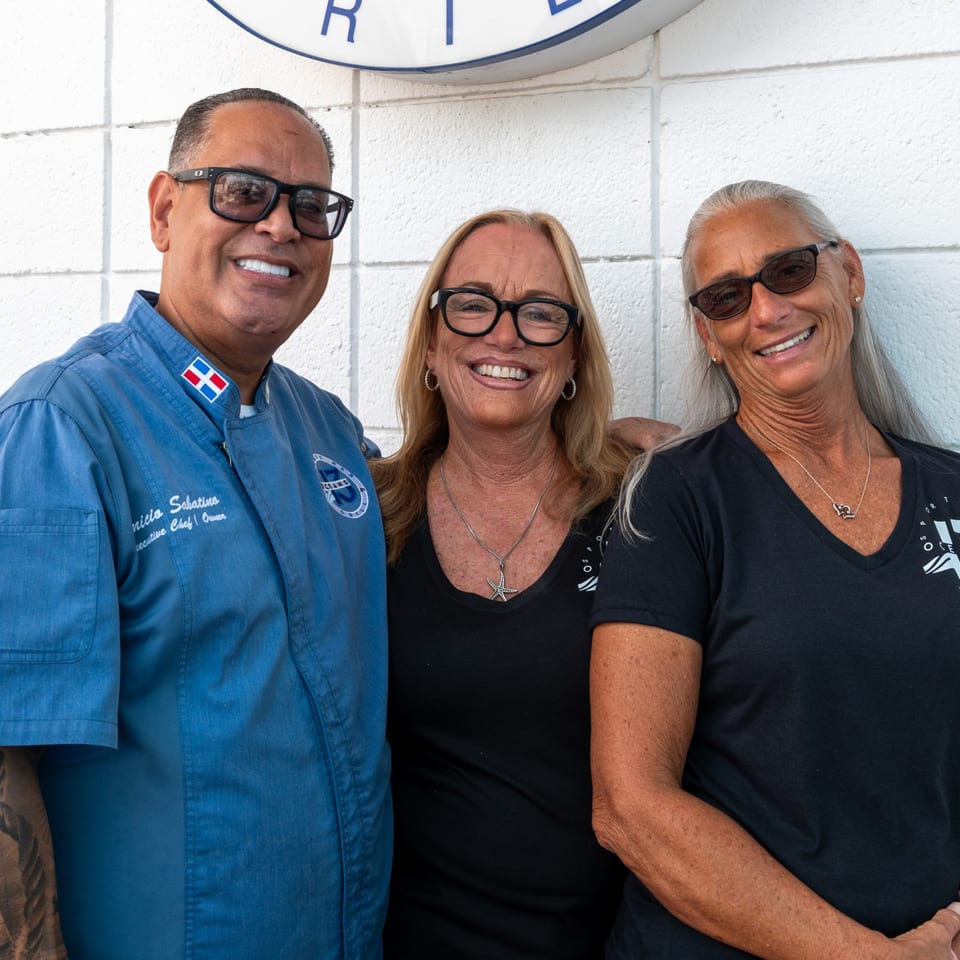 Oceans 13 chef and two women smiling, standing against a white wall with a logo.