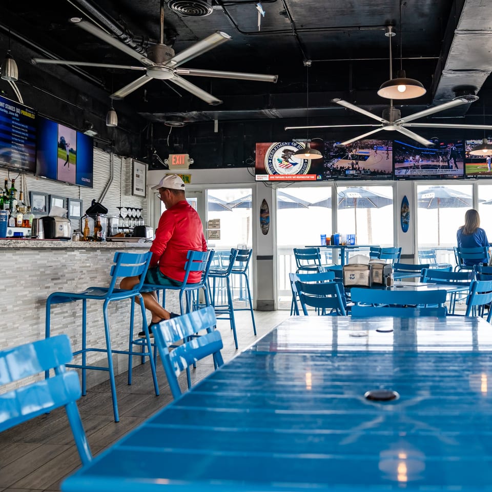 A man in a red shirt sits at a bar with blue stools and tables, watching TV.