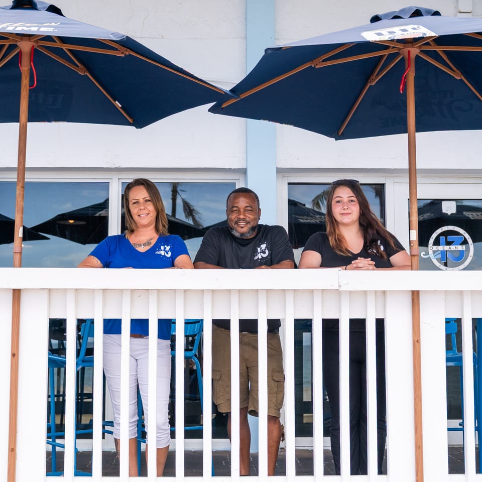 Three Oceans 13 Sports Bar & Grill staff members smiling behind a white railing under blue umbrellas.
