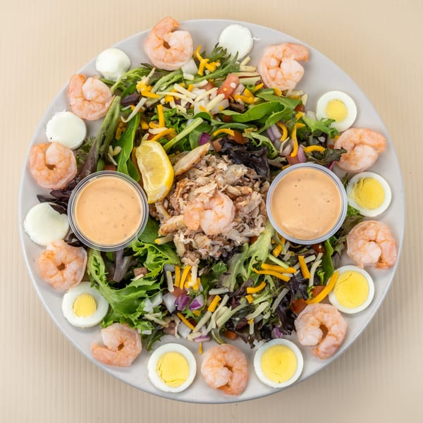 Overhead view of a large salad with shrimp, crab meat, hard-boiled eggs, and two dressing cups.