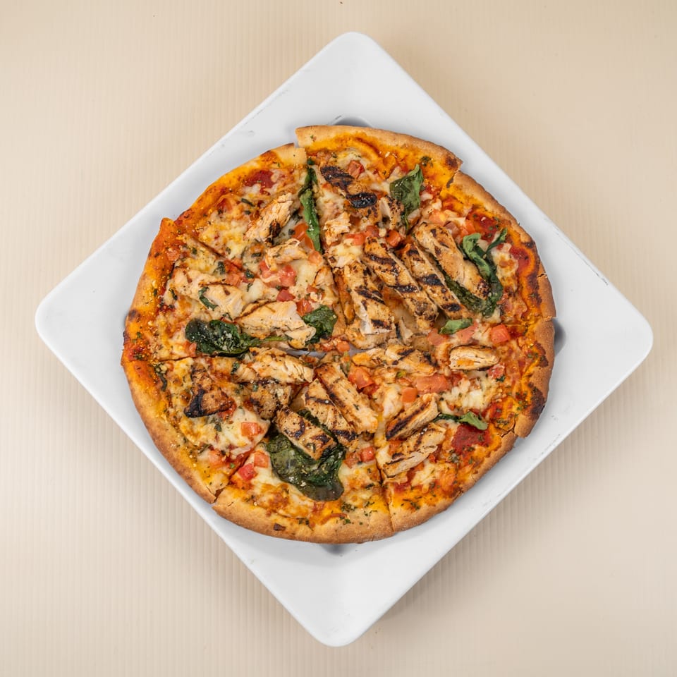 Top-down view of a sliced chicken pizza with tomatoes and spinach on a white square plate.