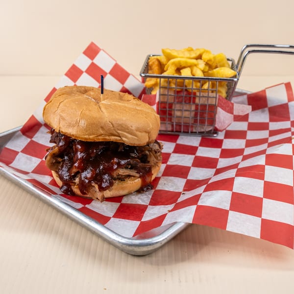 Pulled pork sandwich with BBQ sauce and a side of crinkle-cut fries on a metal tray.