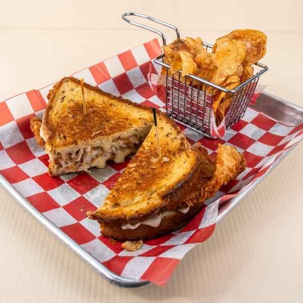 Grilled cheese sandwich with caramelized onions and chips in a metal basket on a red and white checkered tray.