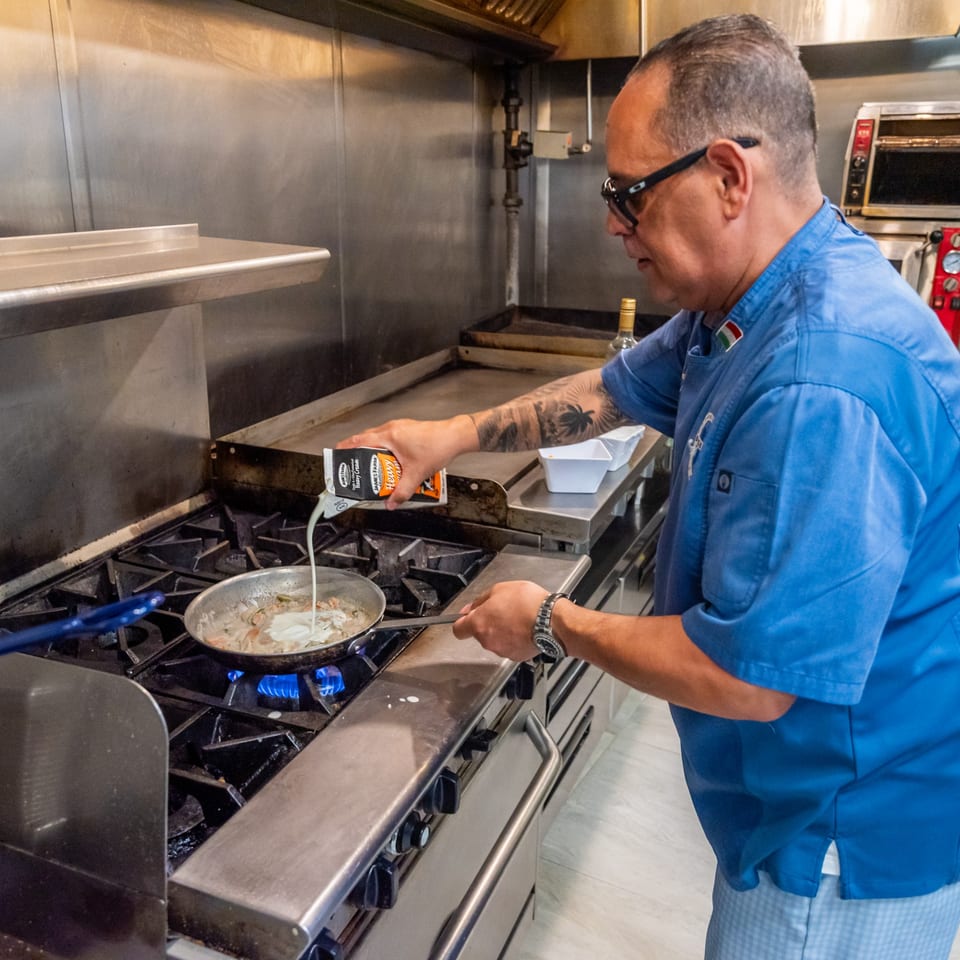 Chef in blue uniform pouring cream into a pan of food cooking on a gas stove.