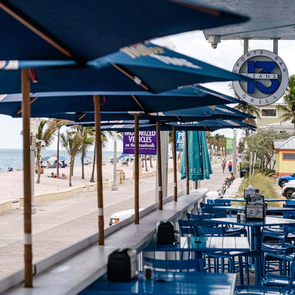 Outdoor dining area with blue tables and umbrellas overlooking a beach with palm trees and ocean.