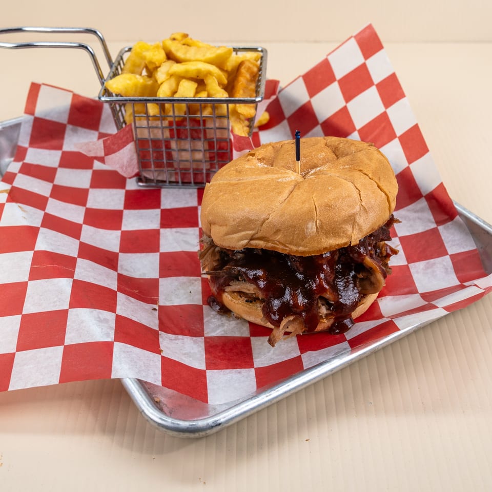 Pulled pork sandwich with BBQ sauce and a side of fries in a metal basket on a red checkered paper.