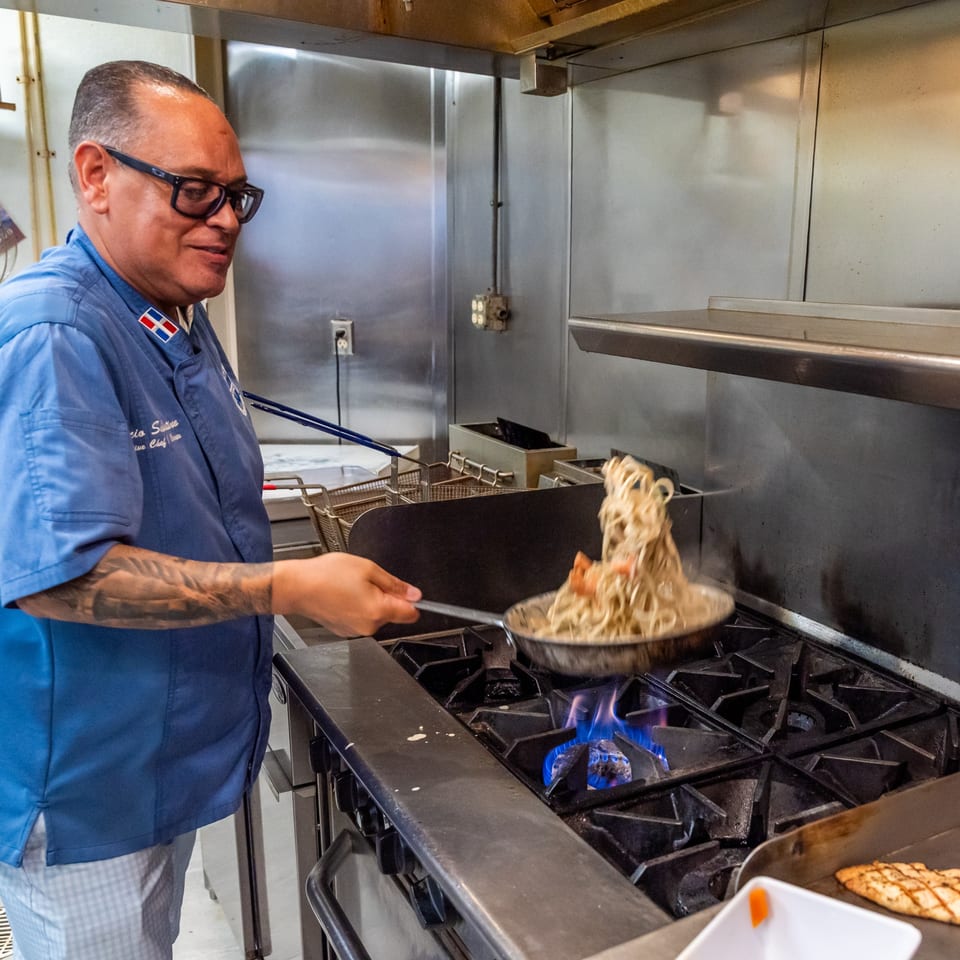 Chef in a blue uniform and glasses cooking pasta in a pan over a gas stove.