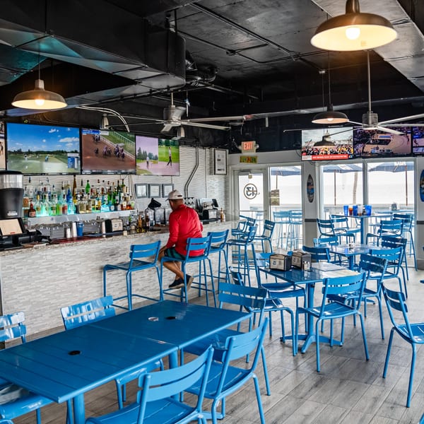 A man sits at a bar with multiple TVs showing sports, surrounded by blue tables and chairs.