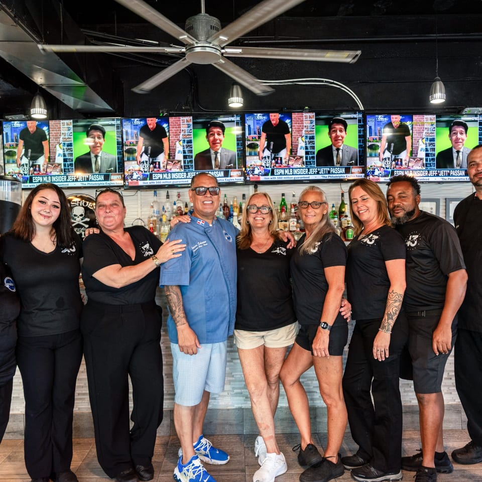 Oceans 13 bar-&-grill staff posing in front of a bar with multiple TVs showing sports.