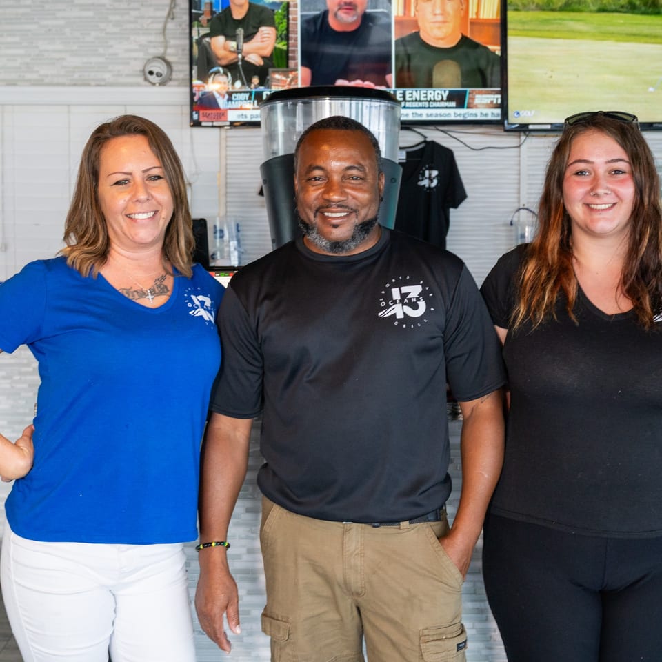 Three smiling restaurant staff members, two women and one man, stand together in front of TVs.