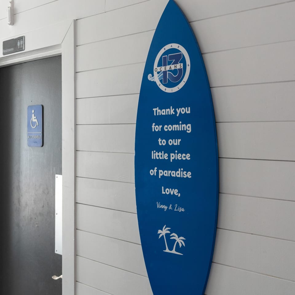 Oceans 13 Bar & Grill surfboard sign with a welcome message next to a restroom door.
