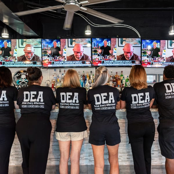 Six Oceans 13 staff in black shirts face a bar with liquor bottles and TVs showing sports.