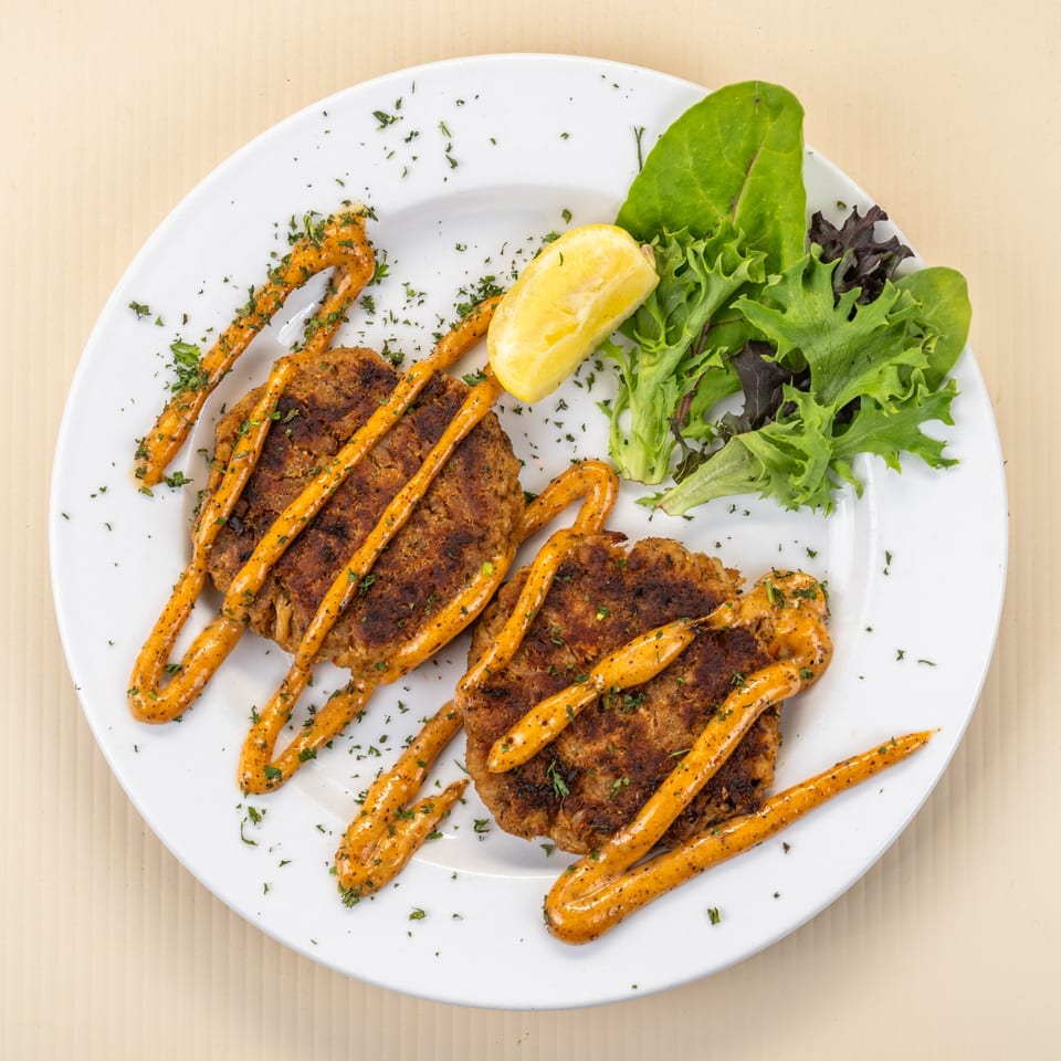 Two crab cakes drizzled with orange sauce, garnished with parsley, lemon, and mixed greens.