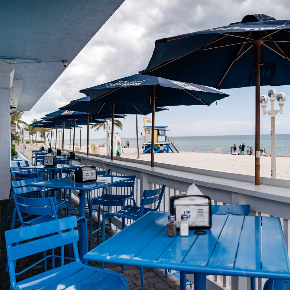 Outdoor dining area with blue tables and chairs, overlooking a sandy beach and ocean with a lifeguard stand.