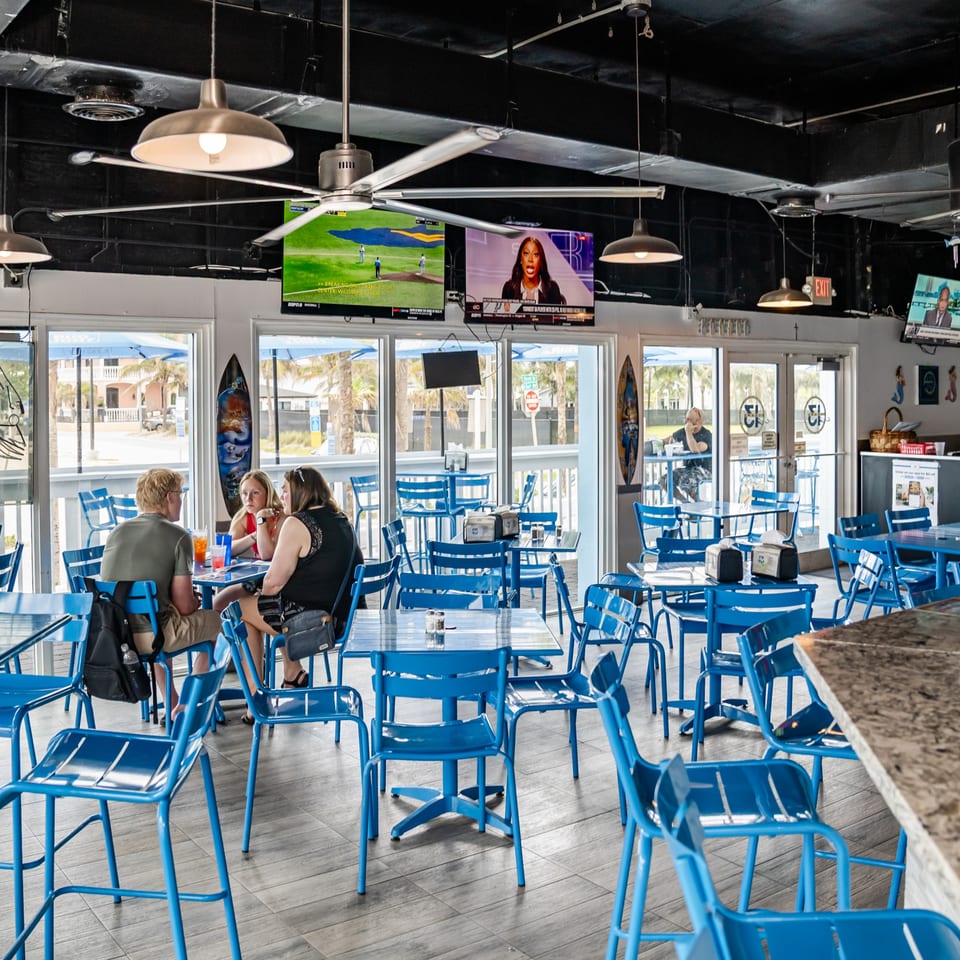 A bright, casual restaurant interior with blue tables and chairs, people dining, and large windows.