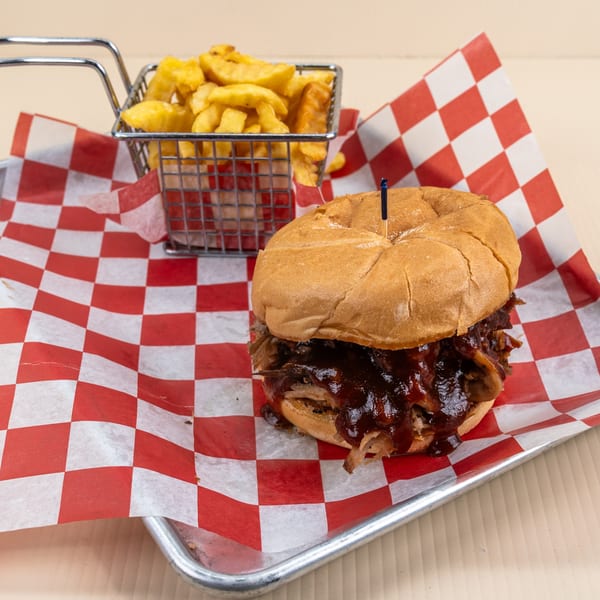 Pulled pork sandwich with barbecue sauce and a side of fries in a metal basket on checkered paper.