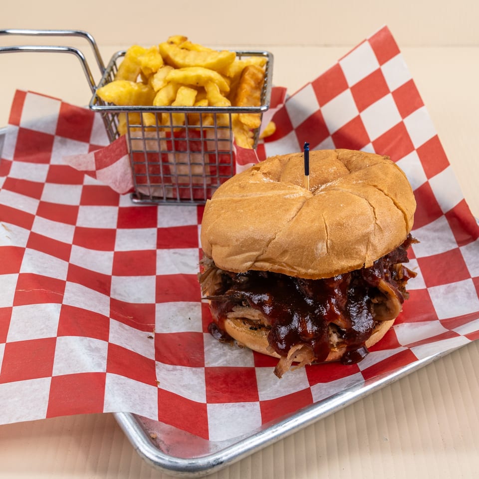Pulled pork sandwich with barbecue sauce and a side of fries in a metal basket on checkered paper.
