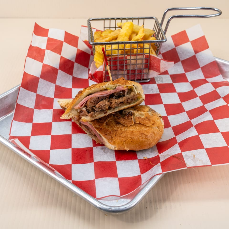 Cuban sandwich cut in half, revealing ham and roasted pork, served with fries on checkered paper.