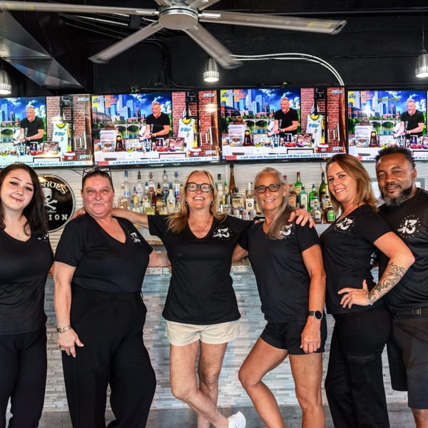 Six Oceans 13 staff members in black shirts smiling behind a bar with liquor bottles and TVs.